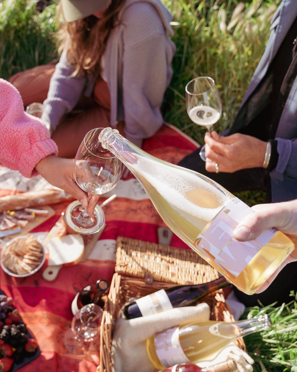 Person pouring bottle of Lily's Pet Nat into glass next to 2 others holding wine glasses over a picnic in the park